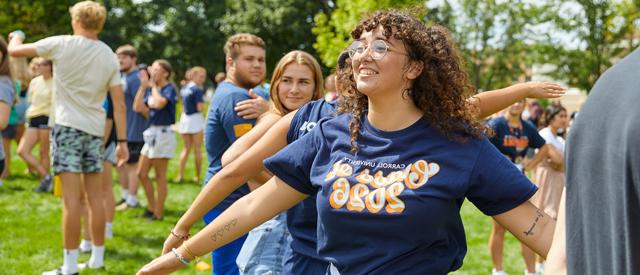 A cheerful young person in a "class of 2026" t-shirt enjoying an outdoor event on a sunny day with other attendees in the background.